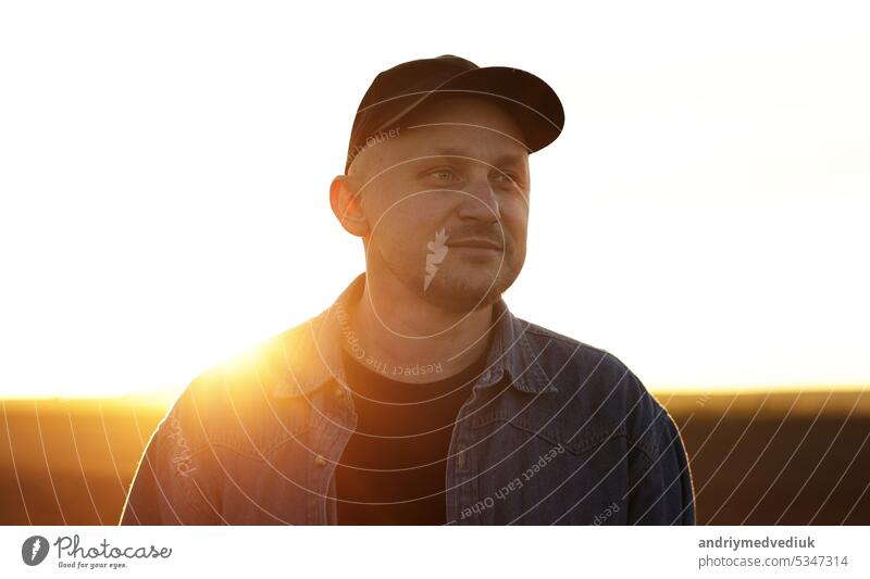 Porträt eines lächelnden männlichen Landwirts mit Mütze, der bei Sonnenuntergang durch ein endloses gepflügtes Ackerfeld geht. Professioneller Agronom inspiziert kultiviertes Land im Sonnenlicht. Agribusiness-Konzept und Landwirtschaft.