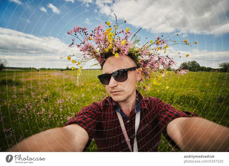 Junger lächelnder Mann mit Wildblumenkranz und Sonnenbrille macht ein Selfie mit einer Weitwinkelkamera und hat Spaß auf dem Feld an einem Sommertag. Positive Kerl hat keine Allergie. Lifestyle Sommer Konzept.
