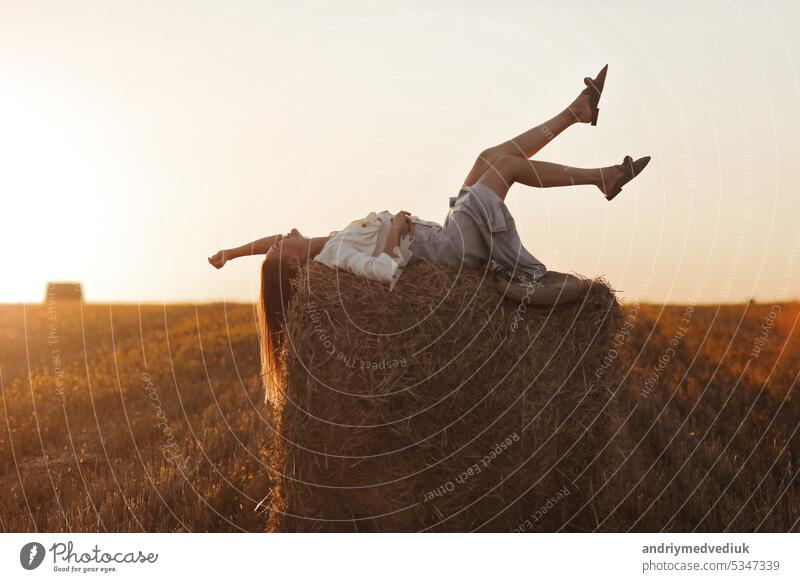 Junge Frau mit langen Haaren, trägt Jeansrock, helles Hemd liegt auf Strohballen im Feld im Sommer bei Sonnenuntergang. Weibliche Porträt in natürlichen ländlichen Szene. Umwelt Ökotourismus Konzept.
