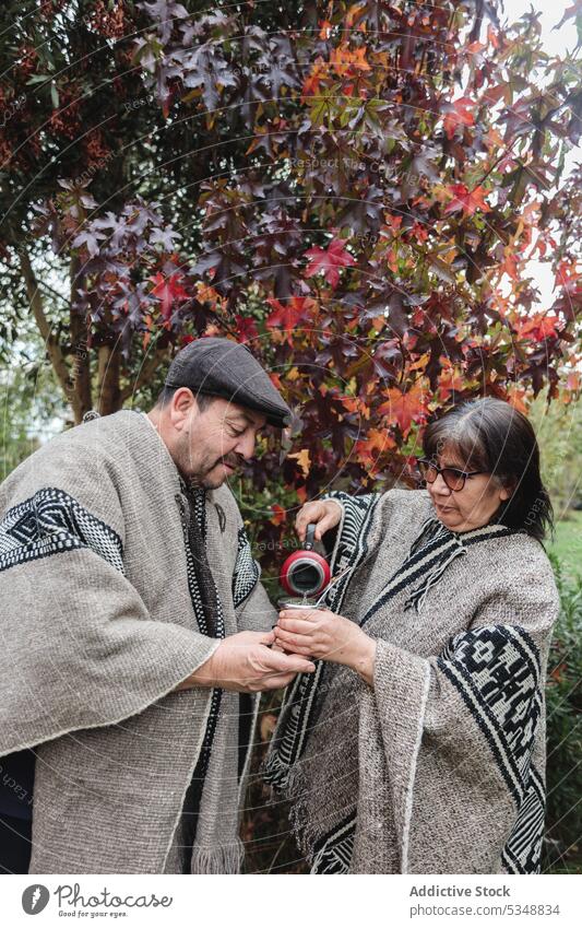 Seriöses reifes Paar in traditioneller Mapuche-Kleidung mit Thermoskanne und Becher gießt heiße Yerba Mate ein, während es im Herbstgarten in Temuco steht