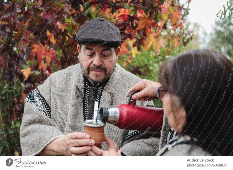 Anonyme Frau serviert einem seriösen Mann heiße Yerba Mate, während sie im Herbstgarten in Temuco steht Paar trinken Yerba Partnerin Inhalt Thermoskanne mapuche
