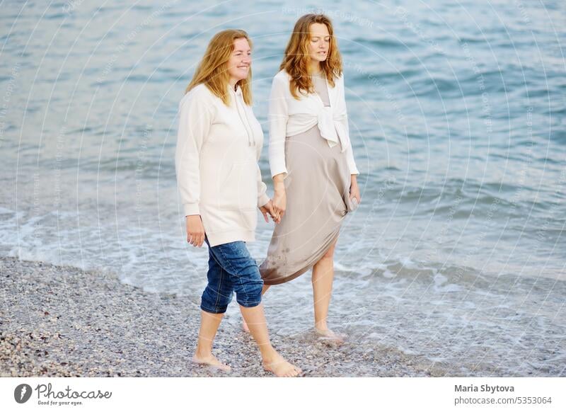 Rothaarige ältere Mutter und ihre erwachsene schöne schwangere Tochter spazieren zusammen am Meeresufer. Senior Erwachsener Menschen Spaziergang Mutterschaft