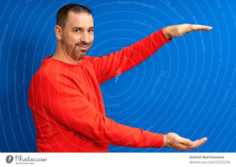 Bärtiger hispanischer Mann in rotem Pullover mit offenen Armen im Profil, der etwas anbietet und in die Kamera lächelt, isoliert auf blauem Hintergrund. Person