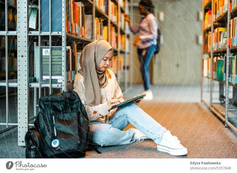 Weibliche muslimische Studentin sitzt in der Bibliothek auf dem Boden und liest mit einem Freund ein Buch echte Menschen Teenager Campus positiv Prüfung Wissen