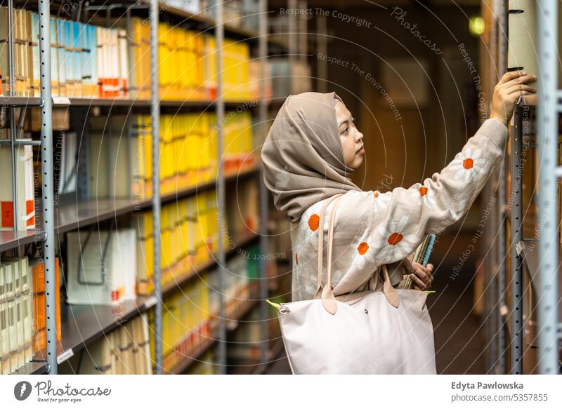 Asiatische muslimische Studentin, die ein Buch aus dem Bücherregal in der Bibliothek nimmt echte Menschen Teenager Campus positiv Prüfung Wissen selbstbewusst
