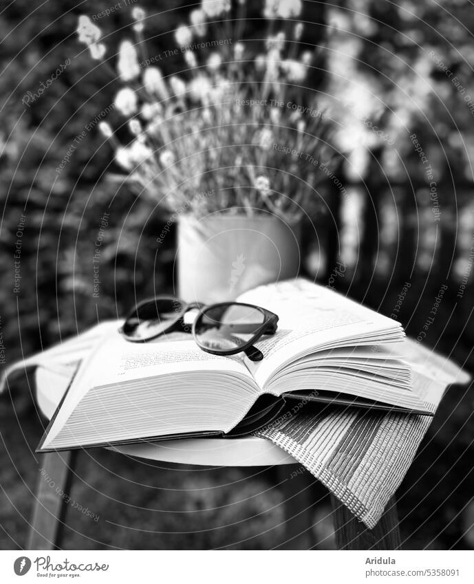 Im Garten liegt auf einem kleinen Tisch ein aufgeschlagenes Buch mit einer schwarzen Brille. Im Hintergrund steht ein weißer Lavendel im Topf s/w Sonnenbrille