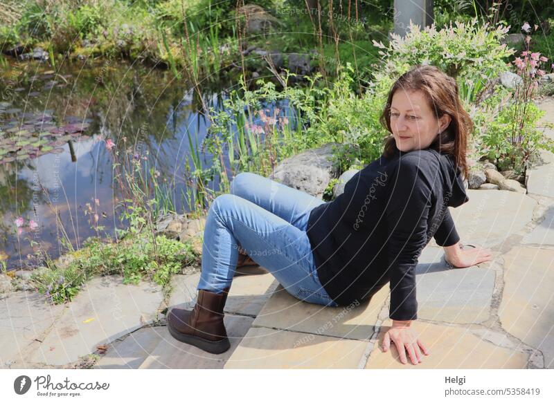 Mainfux-UT | langhaarige Frau sitzt lässig auf Steinstufen am Gartenteich Mensch weiblich feminin Terrasse Stufen Teich Pflanze Blume sitzen Jeans Pulli Shirt