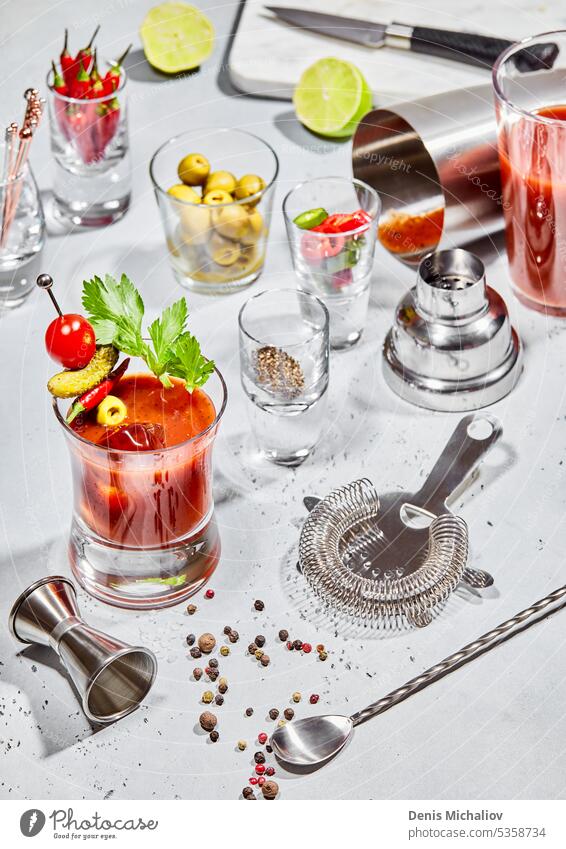 Bloody-Mary-Cocktail-Glas mit Bar-Utensilien, Limette, Paprika, Oliven auf hellem Brett mit Sonnenlicht. blutig Schüttler Vodka Löffel Saft Sieb Tomate Cayenne