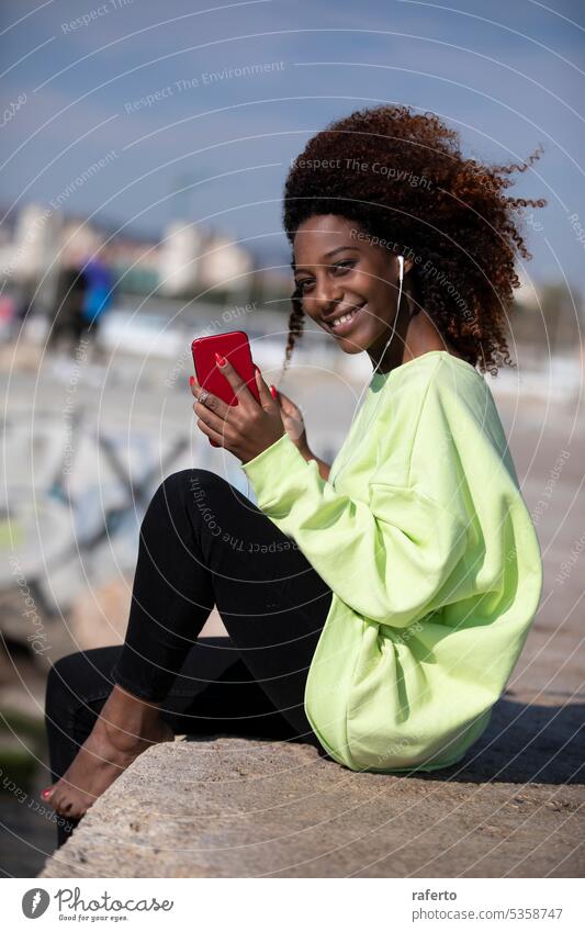 Eine afroamerikanische Frau sitzt am Meer und hört mit ihrem Mobiltelefon in der Hand Musik. Sitzen MEER Musik hören Handy Technik & Technologie Erholung