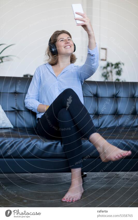 Junge lächelnde Frau sitzt auf einem Ledersofa mit Kopfhörern und macht ein Selfie jung Lächeln Sitzen unter Glück freudig Mädchen Erholung