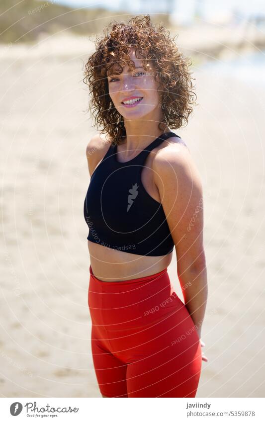 Lächelnde Frau in Sportkleidung am Strand stehend positiv Athlet passen Sportbekleidung Sommer Sonne Hand hinter dem Rücken MEER Glück lockig Leggings Fitness