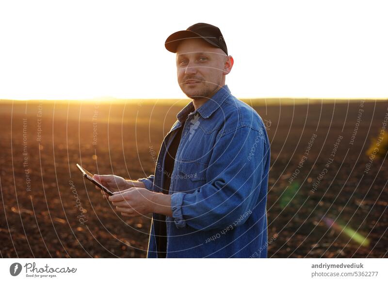 Lächelnder männlicher Landwirt hält ein digitales Tablet auf einem Feld mit gepflügtem Boden bei Sonnenuntergang und schaut in die Kamera. Überprüfung und Kontrolle der Bodenqualität, Vorbereitung des Bodens für die Aussaat von Pflanzen. Intelligente Landwirtschaftstechnologie.