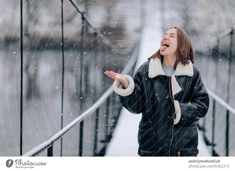 Eine Frau geht und fängt Schneeflocken mit seiner Zunge über den Fluss auf einer Hängebrücke im Winter Tag. Junges Mädchen in warmer Kleidung steht auf einer hölzernen Fußgängerbrücke in kalten verschneiten Tag