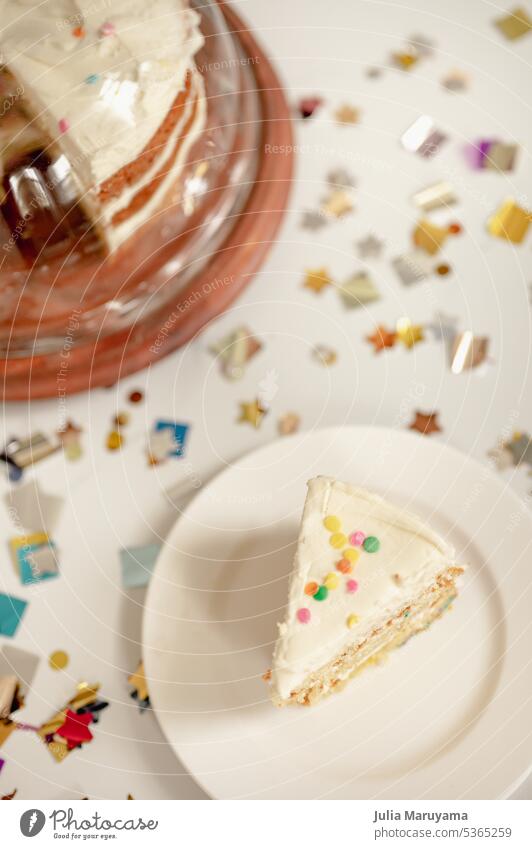 Konfetti-Kuchenscheibe mit bunten Streuseln und geschichtetem Vanillekuchen im überdachten Kuchenständer, umgeben von metallischen Konfetti-Formen