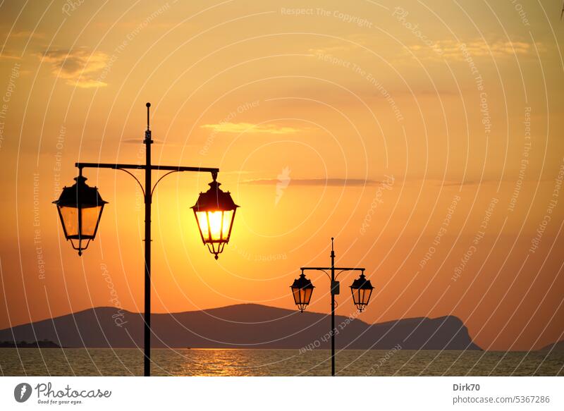 Aufgehende Sonne hinter einer Straßenlaterne mit Blick auf Meer mit Inseln im Hintergrund Mittelmeer Saronischer Golf Ägäis Aegaeisches Meer Laterne
