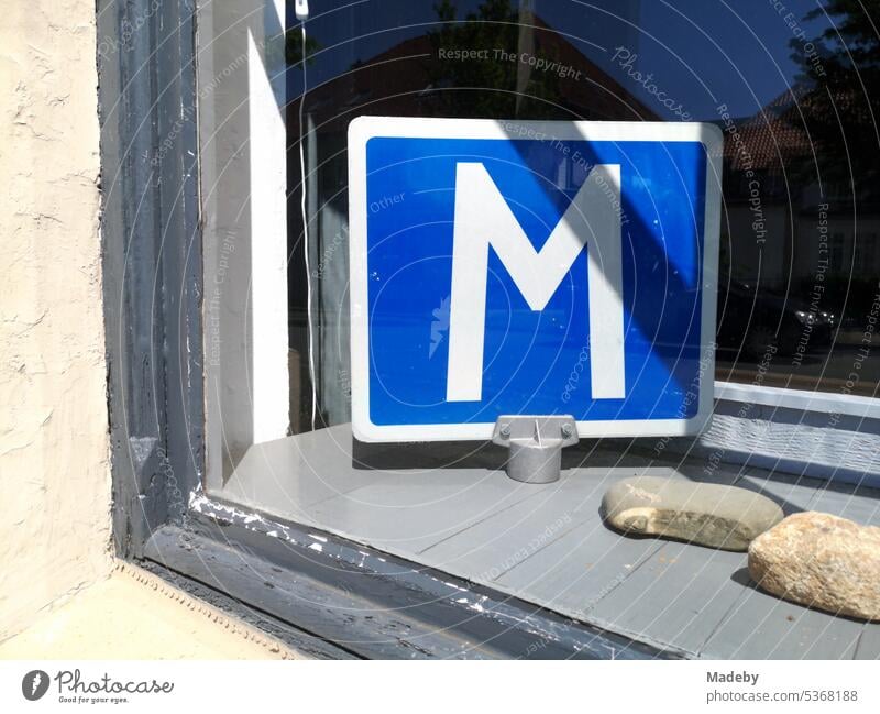 Weißer Buchstabe M auf blauem Grund auf einem Schild im Schaufenster einer Fahrschule im Sommer bei Sonnenschein in Lemgo bei Detmold in Ostwestfalen-Lippe