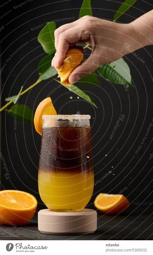 Eiskaffee mit Orangensaft in einem durchsichtigen Glas, die Hand einer Frau drückt eine Orangenscheibe in ein sprudelndes Getränk Ebene Kaffee orange braun süß
