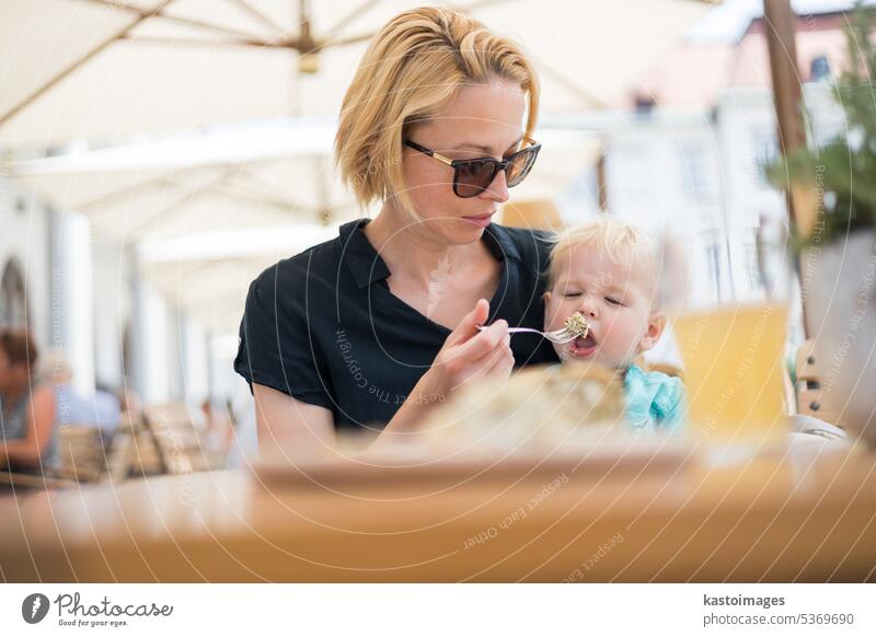 Junge kaukasische blonde Mutter Löffel füttern ihre kleinen Säugling Baby Junge Kind im Freien auf Restaurant oder Café Terrasse im Sommer. Familie Zusammensein