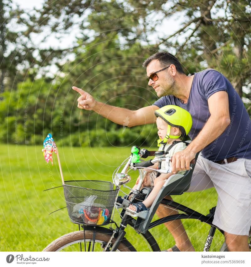 Schauen Sie dort drüben. Aktiver Familientag in der Natur. Vater und Sohn fahren Fahrrad durch den Stadtpark an einem sonnigen Sommertag. Ein niedlicher Junge sitzt im vorderen Fahrradstuhl, während der Vater Fahrrad fährt. Vater-Sohn-Bindung.