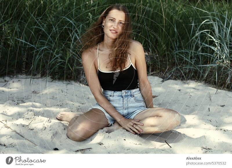 seitliches Portrait einer jungen, schönen, brünetten, lächelnden Frau, die im Sand vor einem Wald kniet warm sommerlich schönes wetter Sommer Pflanzen Nähe Tag