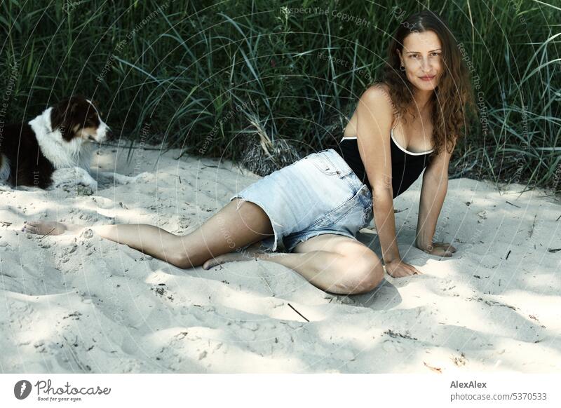 seitliches Portrait einer jungen, schönen, brünetten, lächelnden Frau, die im Sand vor einem Wald sitzt warm sommerlich schönes wetter Sommer Pflanzen Nähe Tag