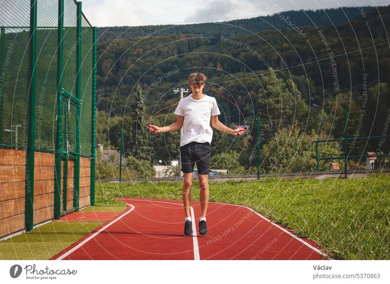 Braunhaariger Junge mit athletischer Figur, der ein weißes T-Shirt und schwarze Shorts trägt, springt Seil auf einem ovalen Sportplatz. Training zur Verbesserung von Sprungkraft, Koordination und Ausdauer