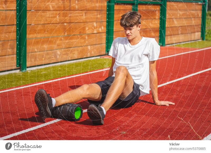 Ein braunhaariger Junge mit athletischem Körperbau auf einem sportlichen Oval massiert seine Muskeln mit einer Schaumstoffrolle, um sich besser zu erholen. Post-Workout