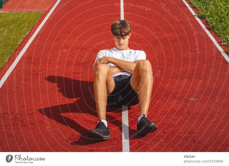 Ein braunhaariger Junge mit athletischem Körperbau auf einem sportlichen Oval massiert seine Muskeln mit einer Schaumstoffrolle, um sich besser zu erholen. Post-Workout