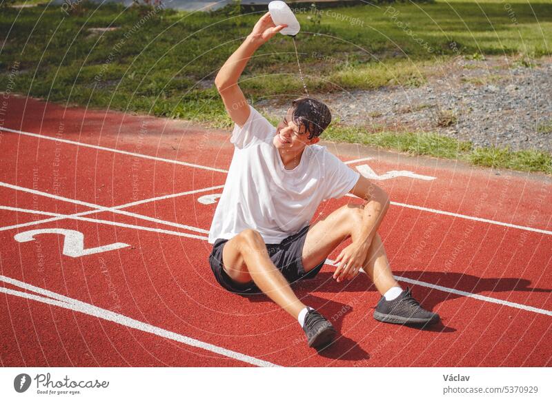 Ein junger Sportler erfrischt sich nach einem harten Training auf dem Leichtathletik-Oval in der großen Hitze mit Wasser. Ausdauertraining. Braunhaariger Jugendlicher