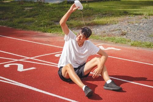 Ein junger Sportler erfrischt sich nach einem harten Training auf dem Leichtathletik-Oval in der großen Hitze mit Wasser. Ausdauertraining. Braunhaariger Jugendlicher
