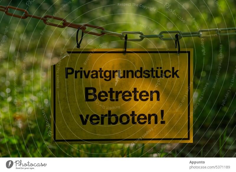 Gelbes Schild "Privatgrundstück - Betreten verboten!" an einer Absperrkette in Nahaufnahme vor einem verschwommenen grünen Hintergrund gelb Verbotsschild privat