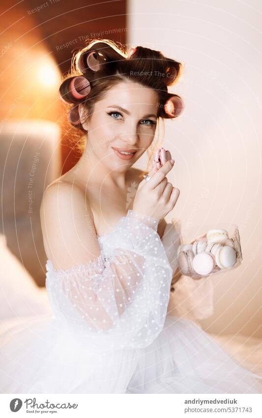 Schönheit Porträt einer schönen lächelnden jungen brünetten Frau mit hellen Pin-up-Make-up essen köstliche Französisch Makronen oder Macarons. Mädchen im weißen Kleid und rosa Lockenwickler sitzen auf dem Bett und entspannen