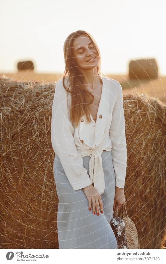Ganzkörperporträt einer lächelnden schönen Brünetten im Jeansrock und mit einer Strohtasche in der Hand. Frau genießt einen Spaziergang in einem Weizenfeld mit Strohballen an einem sonnigen Sommertag.