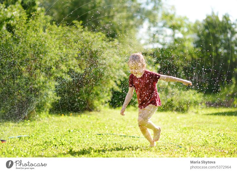 Lustiger kleiner Junge spielt mit Garten Sprinkler in sonnigen Hinterhof. Vorschulkind lacht, springt und hat Spaß mit Sprühwasser. Kind Sprinkleranlage spielen