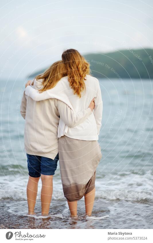 Rothaarige ältere Mutter und ihre erwachsene schöne Tochter spazieren zusammen am Meeresufer. Senior Umarmen vertikal Erwachsener Rücken unkenntlich Natur