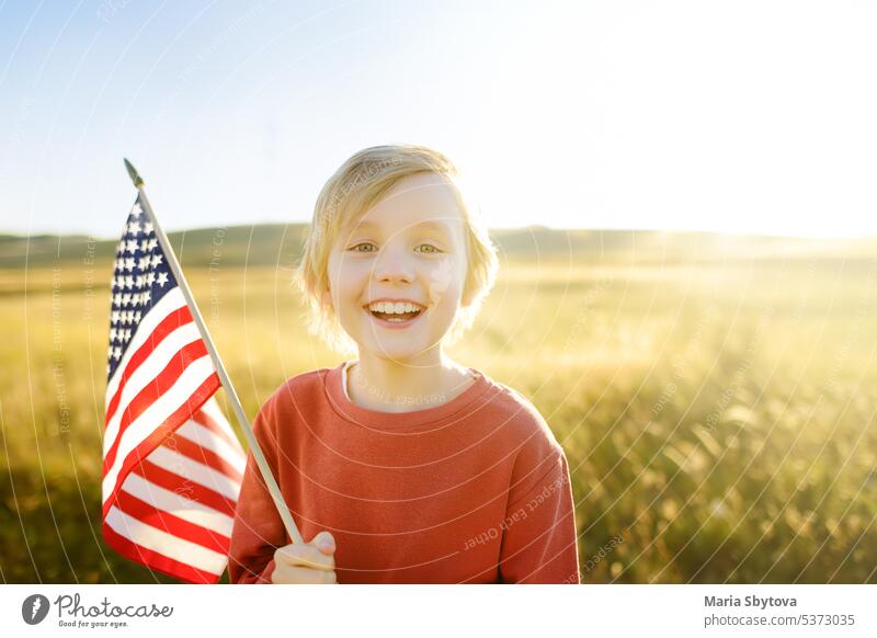Niedlicher kleiner Junge, der den 4. Juli, den Unabhängigkeitstag der USA, bei sonnigem Sommer-Sonnenuntergang feiert. Kind läuft mit amerikanischer Flagge der Vereinigten Staaten auf Weizenfeld. Stolzer kleiner amerikanischer Junge hält Landesflagge