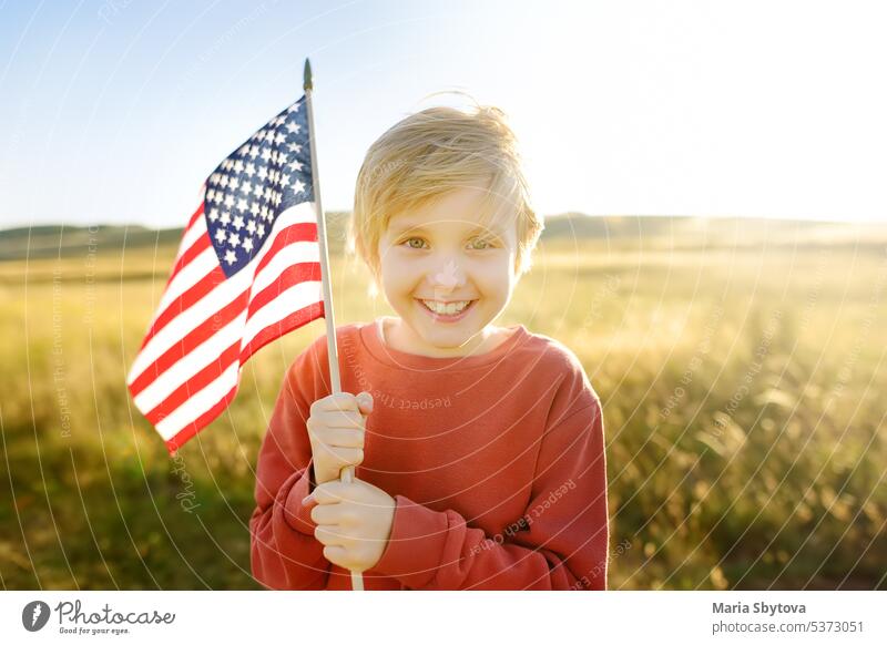 Niedlicher kleiner Junge, der den 4. Juli, den Unabhängigkeitstag der USA, bei sonnigem Sommer-Sonnenuntergang feiert. Kind läuft mit amerikanischer Flagge der Vereinigten Staaten auf Weizenfeld. Stolzer kleiner amerikanischer Junge hält Landesflagge