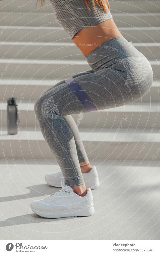 Sportliche, athletische Frau in grauer Sportkleidung macht Übungen mit gymnastischen Gummibändern, Sit-ups im Freien Training Körper Gesäß Kniebeuge Beine