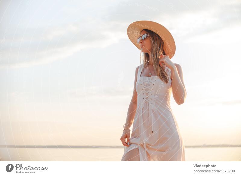Junge kaukasische Frau in weißem Kleid, Sommerhut und Sonnenbrille posiert am Strand bei Sonnenuntergang Hut MEER attraktiv Lächeln posierend Schiff Schwingung