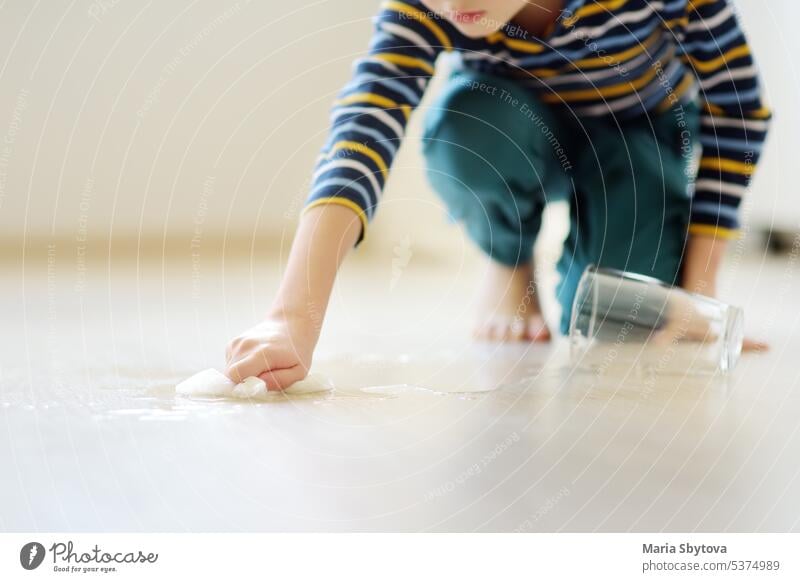 Kleiner Junge wischt verschüttetes Wasser aus einem Glas auf dem Boden auf. Einem Kind beibringen, hinter sich aufzuräumen. Verantwortung, Genauigkeit. Hilfe im Haushalt. Pflichten im Haushalt.