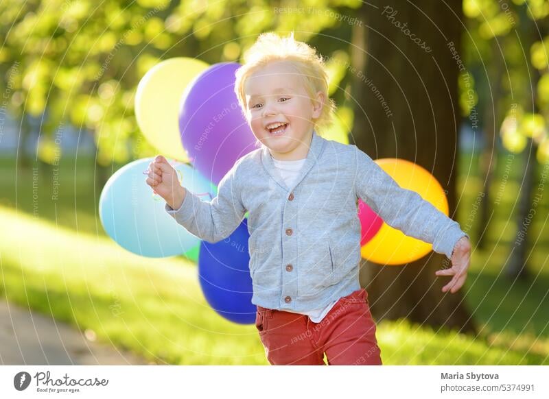 Kleiner Junge mit Spaß während der Feier Geburtstagsparty. Glückliches Kind mit einem Bündel von hellen multicolor Luftballons. Vorschüler oder Kleinkinder Baby-Geburtstagsparty in sonnigen Park. Sommerfest im Freien