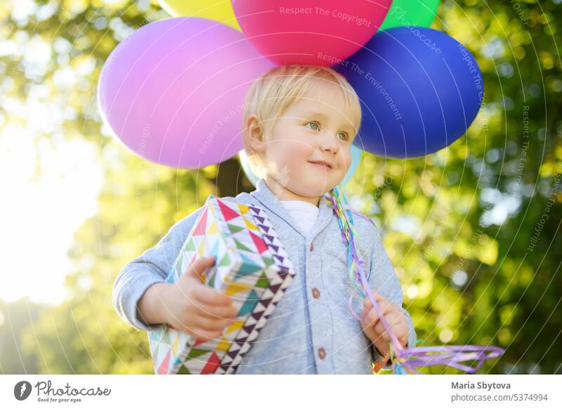 Nettes kleines Kind, das einem Freund zum Geburtstag gratuliert. Kleinkind hält Bündel von bunten Luftballons und Geschenk in einer festlichen Box. Vorfreude auf den Urlaub.