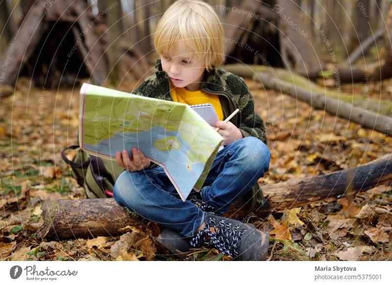 Kleiner Pfadfinder ist Orientierungslauf im Wald. Das Kind sitzt auf einem umgestürzten Baum und schaut auf die Karte im Hintergrund der Tipi-Hütte. wandern