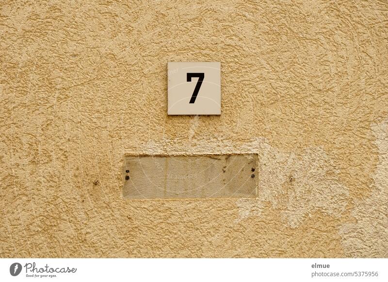 kleines Schild mit einer 7 unter dem ein weiteres Schild optisch sichtbar entfernt wurde an einer verputzten beigefarbenen Hauswand zu vermieten Nummer 7