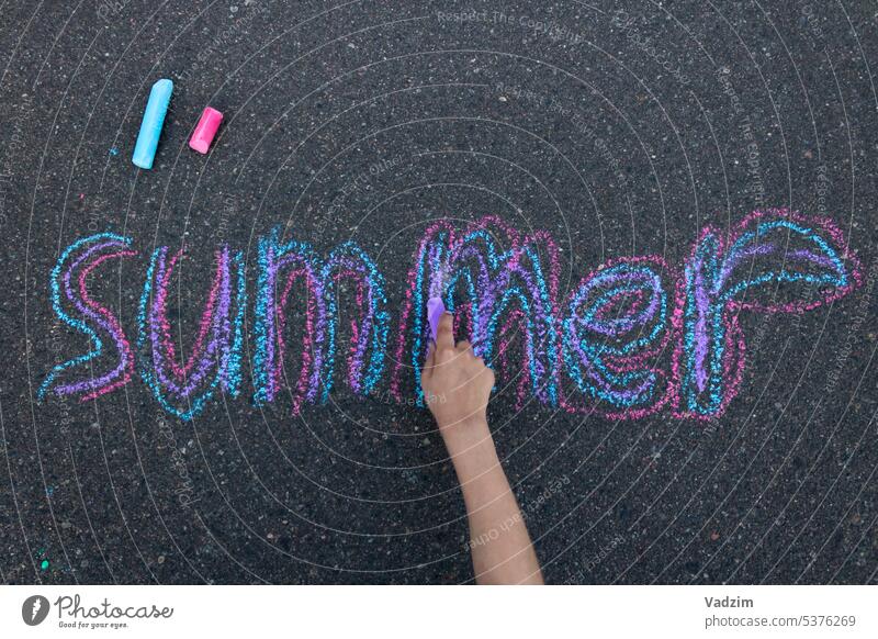 Das Kind schreibt das Wort Sommer mit bunter Kreide auf den Bürgersteig. Ansicht von oben auf die Hand. Asphalt farbenfroh Buntstifte Kindheit Zeichnung Straße
