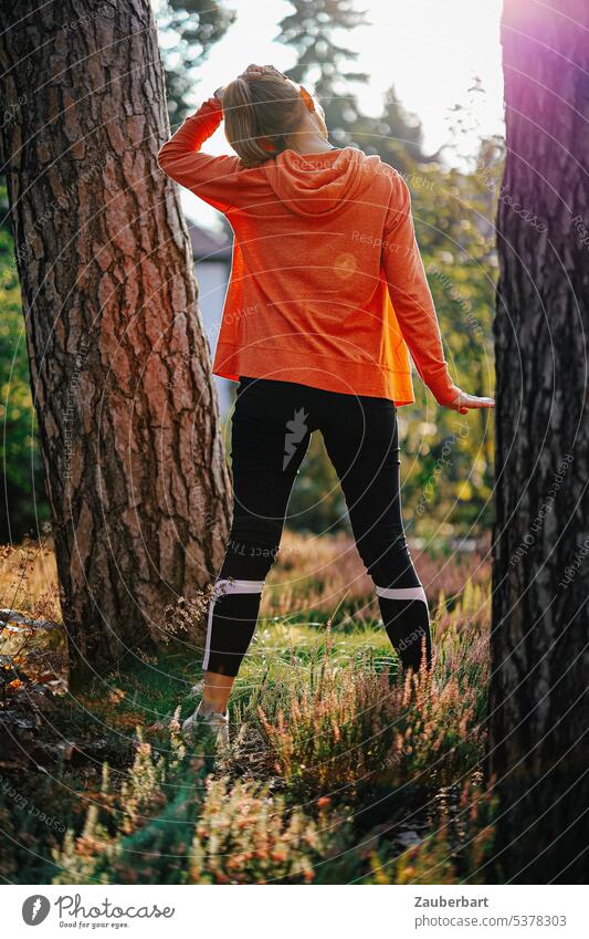 Frau beim Joggen in Rückenansicht bei Dehnübung zwischen zwei Bäumen joggen Sport laufen Dehnen Wald Natur orange Gegenlicht Fitness sportlich Gesundheit