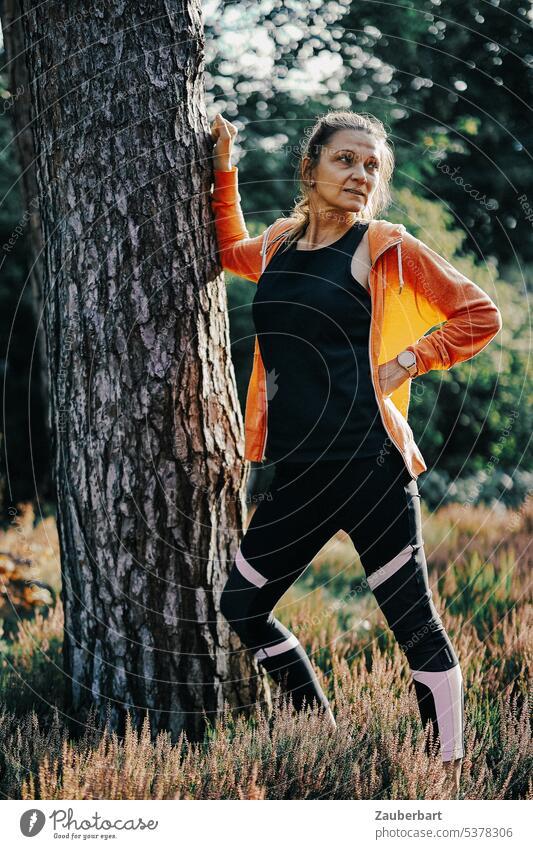 Frau beim Joggen im Wald bei Dehnübung am Baum joggen Sport laufen Dehnen Natur orange Gegenlicht Fitness sportlich Gesundheit Training Lifestyle Läufer rennen