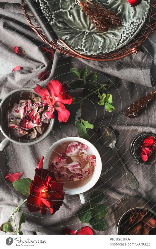Süßer und köstlicher Rosentee in Tassen auf drapiertem grauem Stoff mit Rosen und Blütenblättern. Ansicht von oben Roséwein Tee Becher süß lecker Gewebe