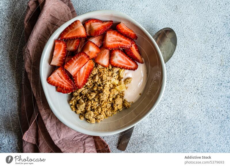 Draufsicht auf eine Schale mit Müsli, Joghurt und reifen Bio-Erdbeeren auf grauer Fläche mit Löffel und Serviette Hintergrund Beeren Schalen & Schüsseln