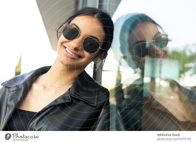 Fröhliche Frau mit Sonnenbrille und Lederjacke Lächeln Glück Straße Stil Großstadt urban Vorschein Mode positiv Outfit trendy Fenster modern Gebäude